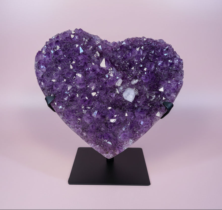 Heart-shaped amethyst crystal cluster on black stand against light pink background