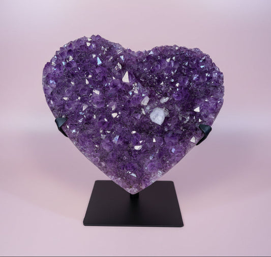 Heart-shaped amethyst crystal cluster on black stand against light pink background