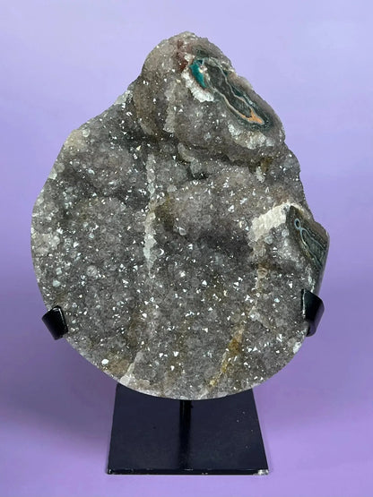 Moody gray rainbow amethyst crystal with druzy, displayed on a black stand against purple background